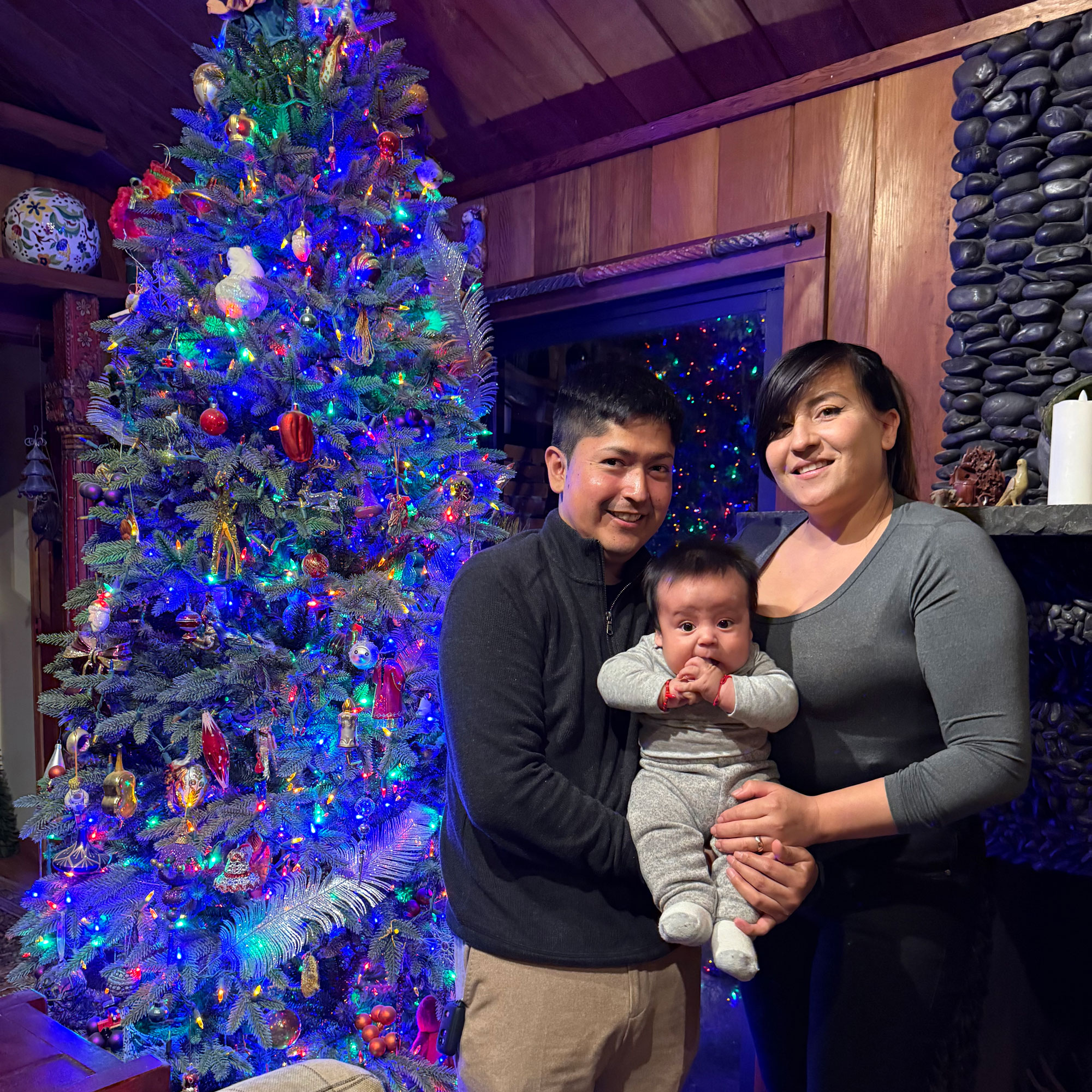 Karla and Birhon holding Gael beside a colorful Christmas tree.